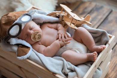 Cute newborn baby wearing aviator hat with toy sleeping in wooden crate