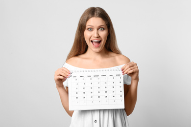 Young woman holding calendar with marked menstrual cycle days on light background