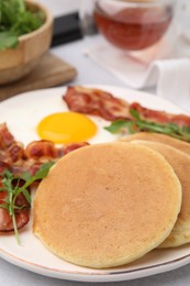 Plate with tasty pancakes, fried egg, arugula and bacon on light table, closeup