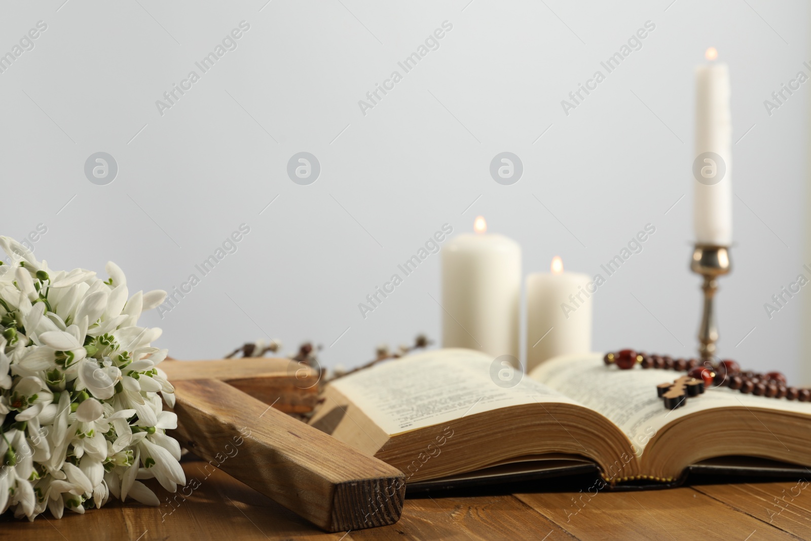 Photo of Church candles, cross, rosary beads, Bible and flowers on wooden table against light background