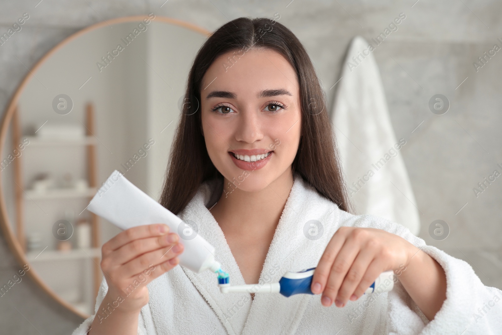 Photo of Young woman squeezing toothpaste from tube onto electric toothbrush in bathroom