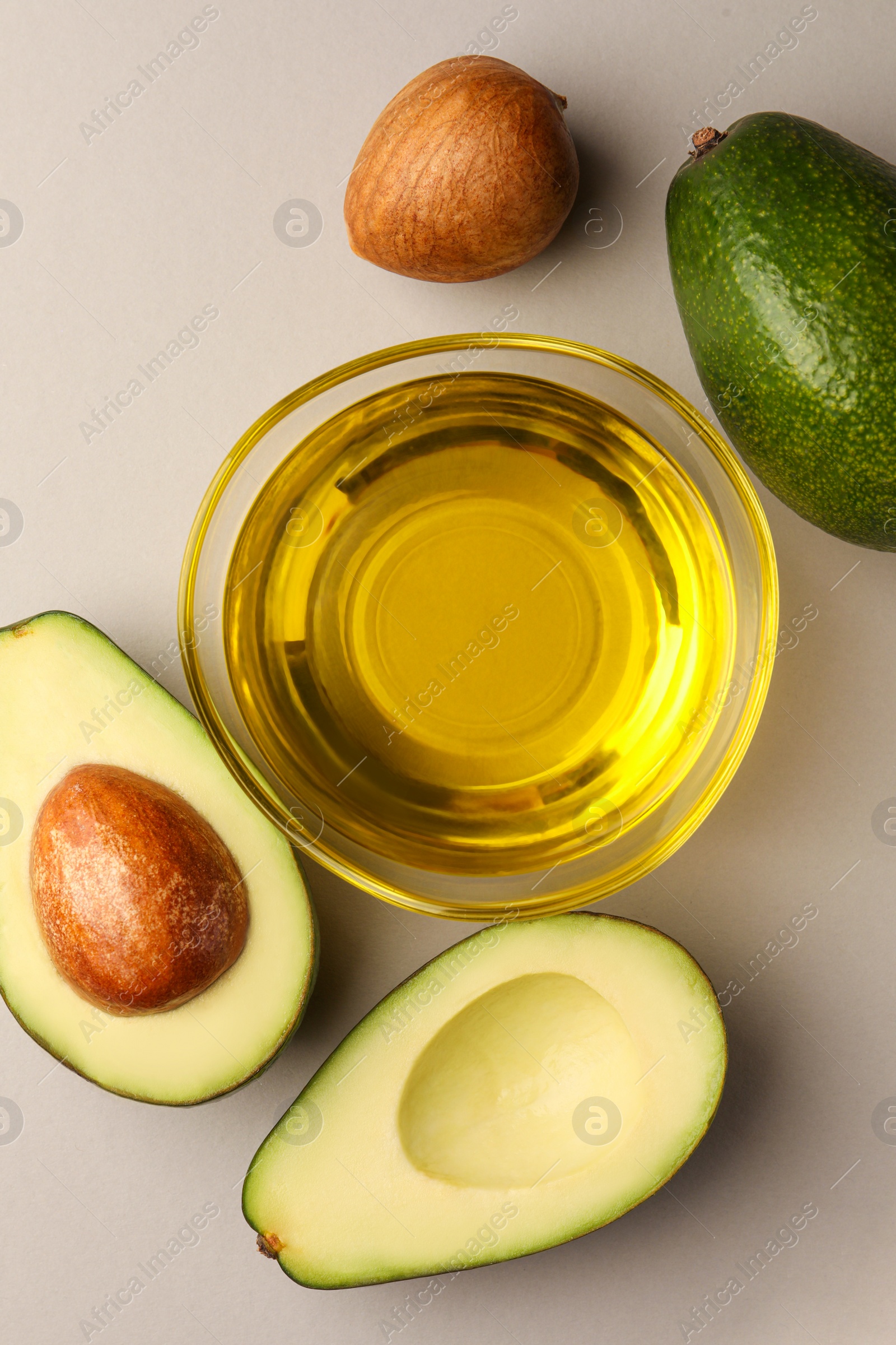 Photo of Cooking oil in bowl and fresh avocados on light grey background, flat lay