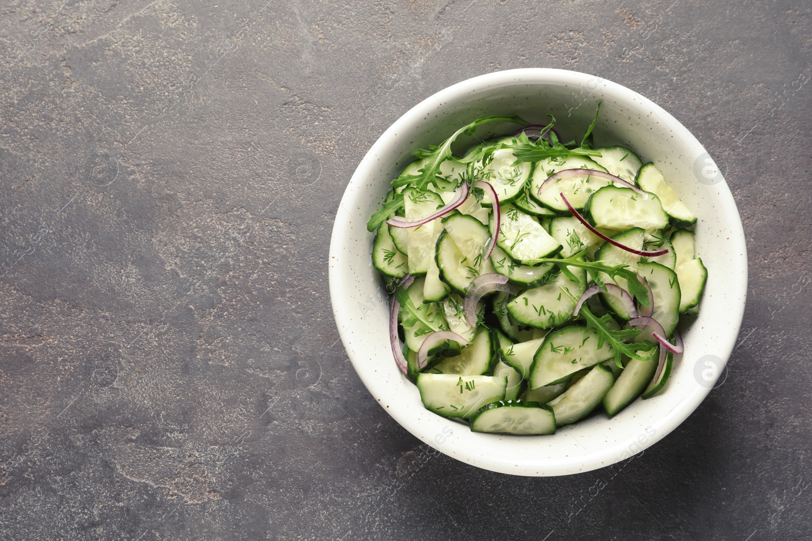 Photo of Delicious cucumber salad with onion and arugula in bowl on grey background, top view. Space for text