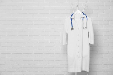 Photo of Medical uniform and stethoscope hanging on rack near white brick wall. Space for text