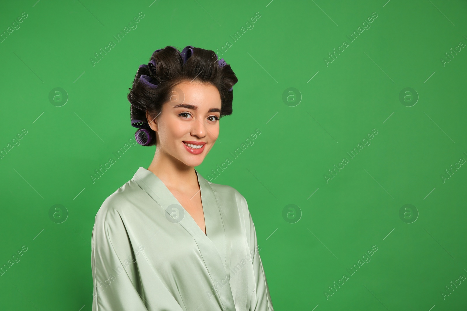 Photo of Happy young woman in silk bathrobe with hair curlers on green background, space for text