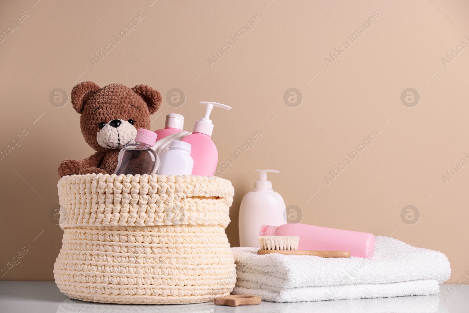 Photo of Knitted basket with baby cosmetic products, bath accessories and toy bear on white table against beige background