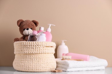 Knitted basket with baby cosmetic products, bath accessories and toy bear on white table against beige background