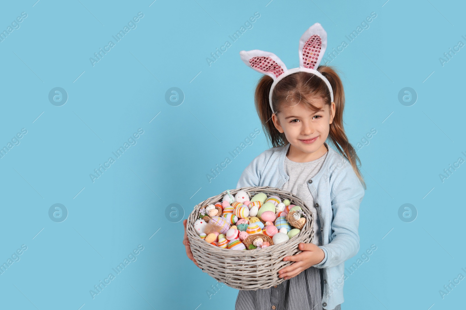 Photo of Happy little girl with bunny ears holding wicker basket full of Easter eggs on light blue background. Space for text