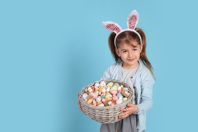 Photo of Happy little girl with bunny ears holding wicker basket full of Easter eggs on light blue background. Space for text