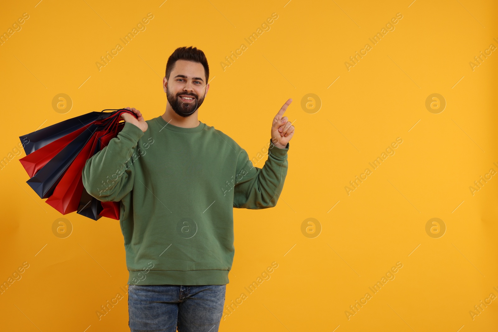 Photo of Happy man with many paper shopping bags pointing at something on orange background. Space for text