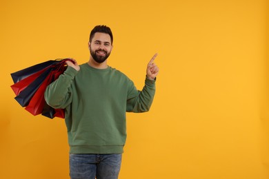 Happy man with many paper shopping bags pointing at something on orange background. Space for text