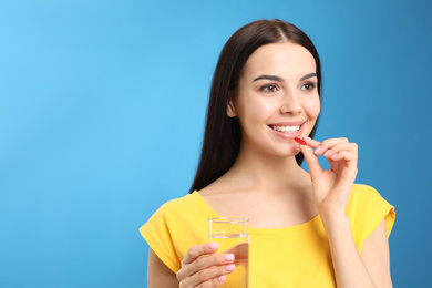 Young woman with glass of water taking vitamin capsule on light blue background. Space for text