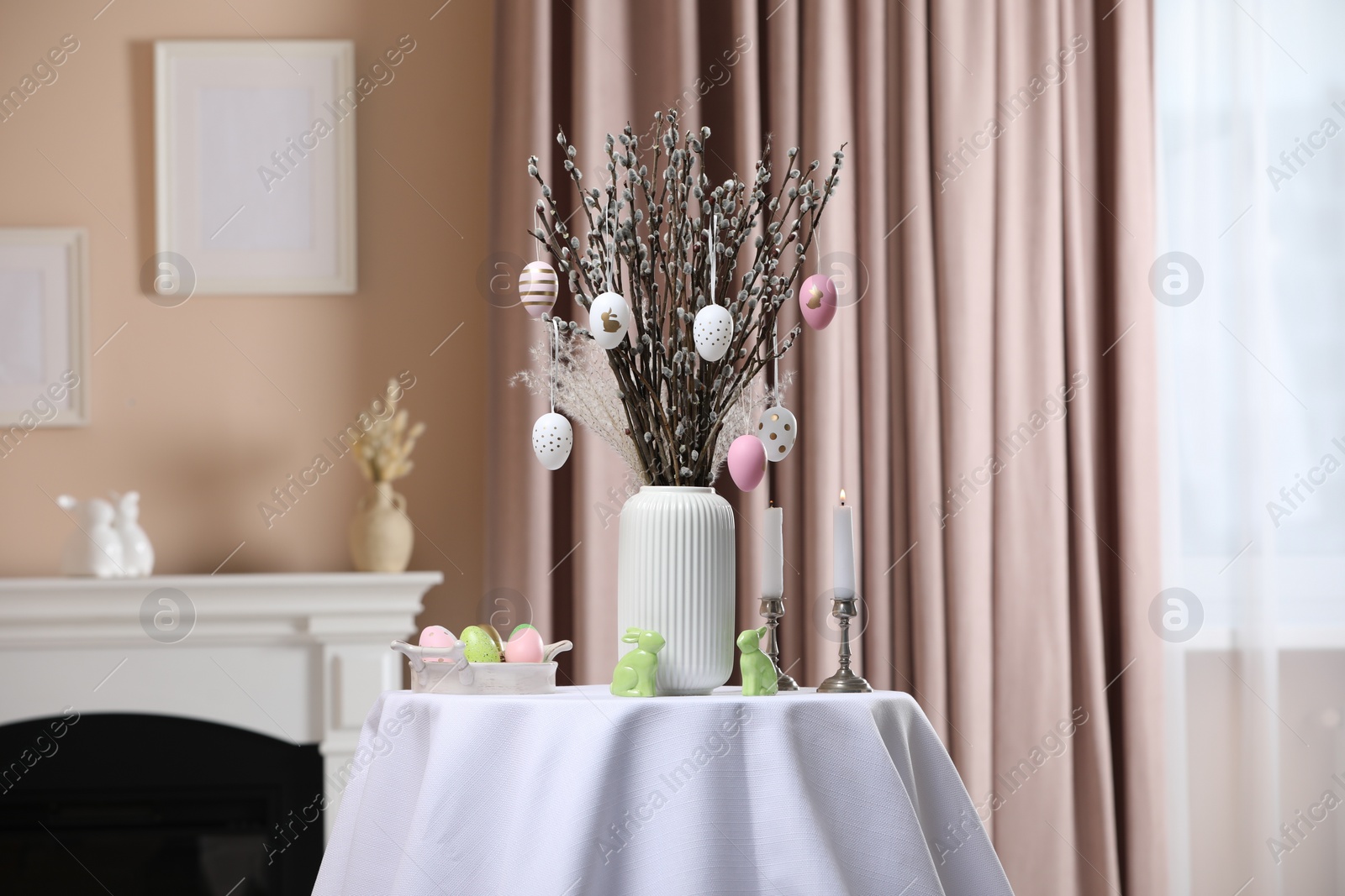 Photo of Pussy willow branches with festively decorated eggs, Easter bunnies and candles on table indoors