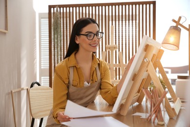 Photo of Young woman drawing on easel at table indoors