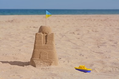 Photo of Beautiful sand castle with small Ukrainian flag and toy boat on beach near sea, space for text