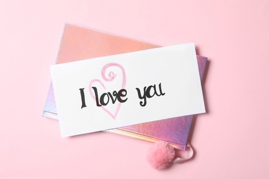 Photo of Card with text I Love You and shiny notebook on pink background, top view