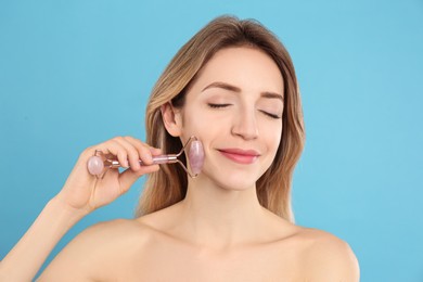 Young woman using natural rose quartz face roller on light blue background
