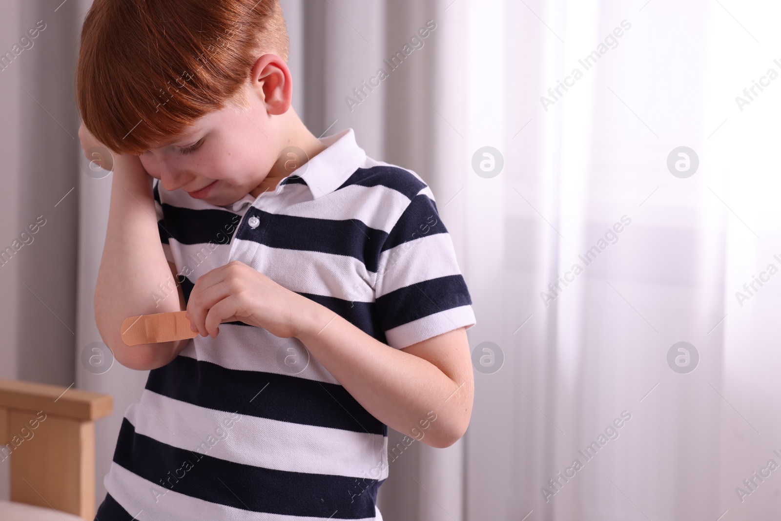 Photo of Little boy putting sticking plaster onto elbow indoors. Space for text