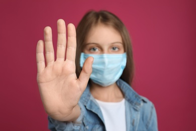 Photo of Little girl in protective mask showing stop gesture on crimson background. Prevent spreading of coronavirus