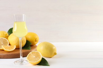 Photo of Liqueur glass with tasty limoncello, lemons and green leaves on white wooden table. Space for text
