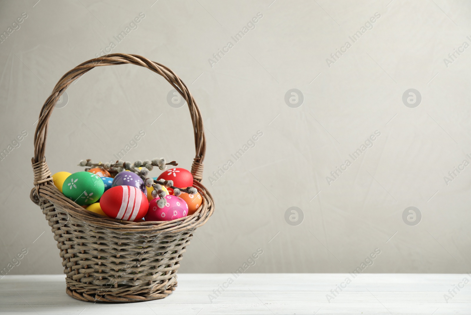 Photo of Colorful Easter eggs in wicker basket on white wooden table against grey background