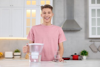 Photo of Happy man with filter jug and glass of clear water at table in kitchen