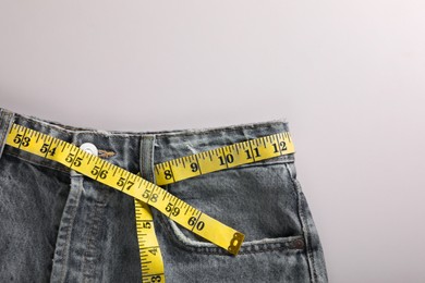 Jeans with measuring tape on light grey background, top view and space for text. Weight loss concept