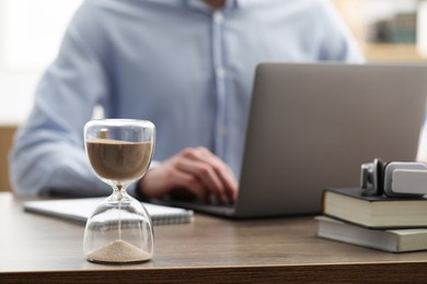 Photo of Hourglass with flowing sand on desk. Man using laptop indoors, selective focus