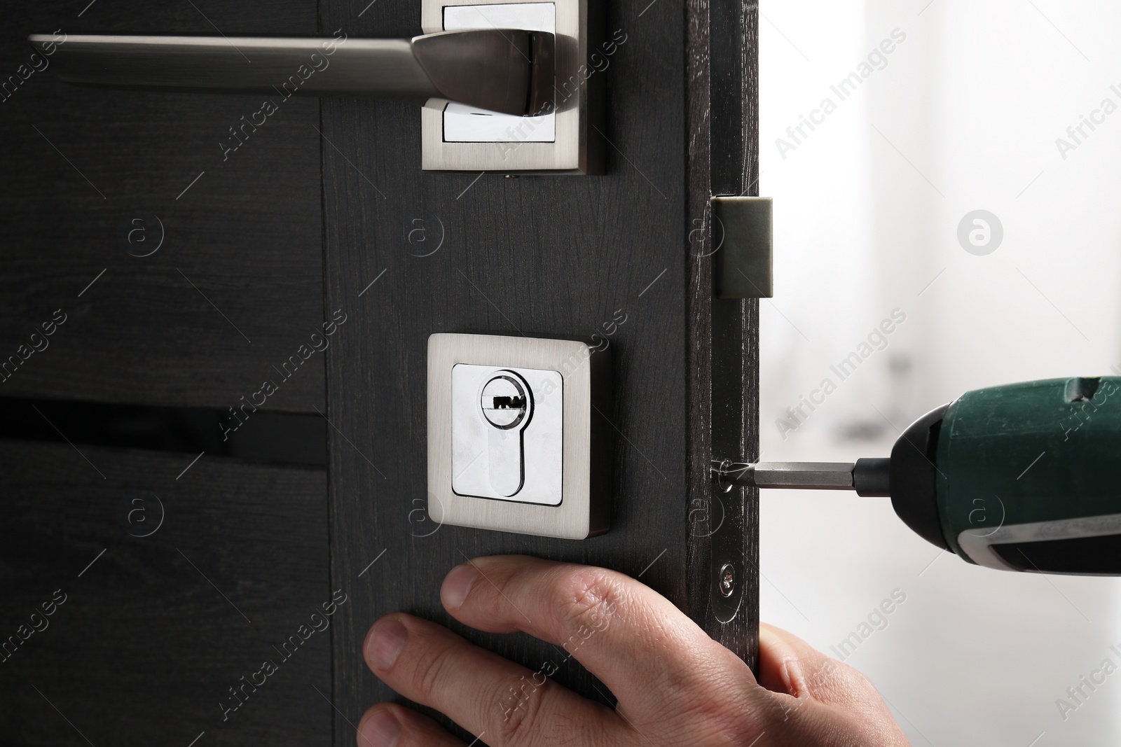 Photo of Handyman with electric screwdriver repairing door handle indoors, closeup