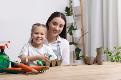 Photo of Mother with her daughter spraying water onto vegetable seeds in peat pots at wooden table indoors