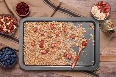 Photo of Making granola. Baking tray with mixture of oat flakes, other ingredients and spoon on wooden table, flat lay