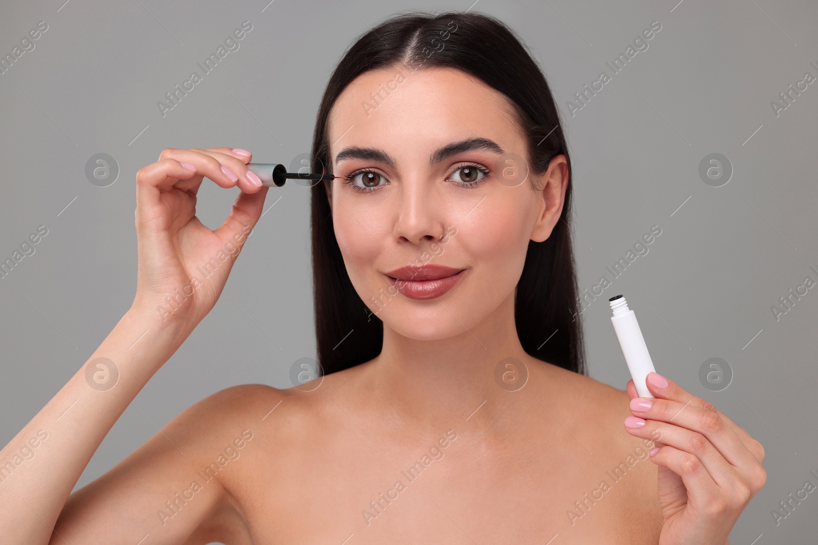 Photo of Beautiful woman applying serum onto her eyelashes on grey background. Cosmetic product