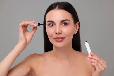 Beautiful woman applying serum onto her eyelashes on grey background. Cosmetic product