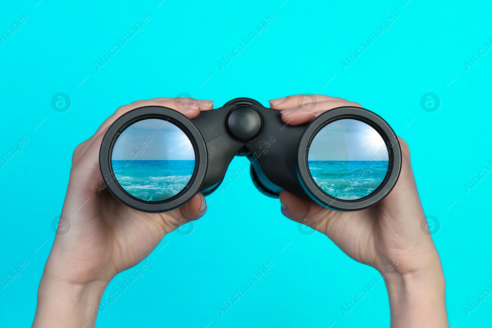 Image of Woman holding modern binoculars on light blue background, closeup. Seascape reflecting in lenses