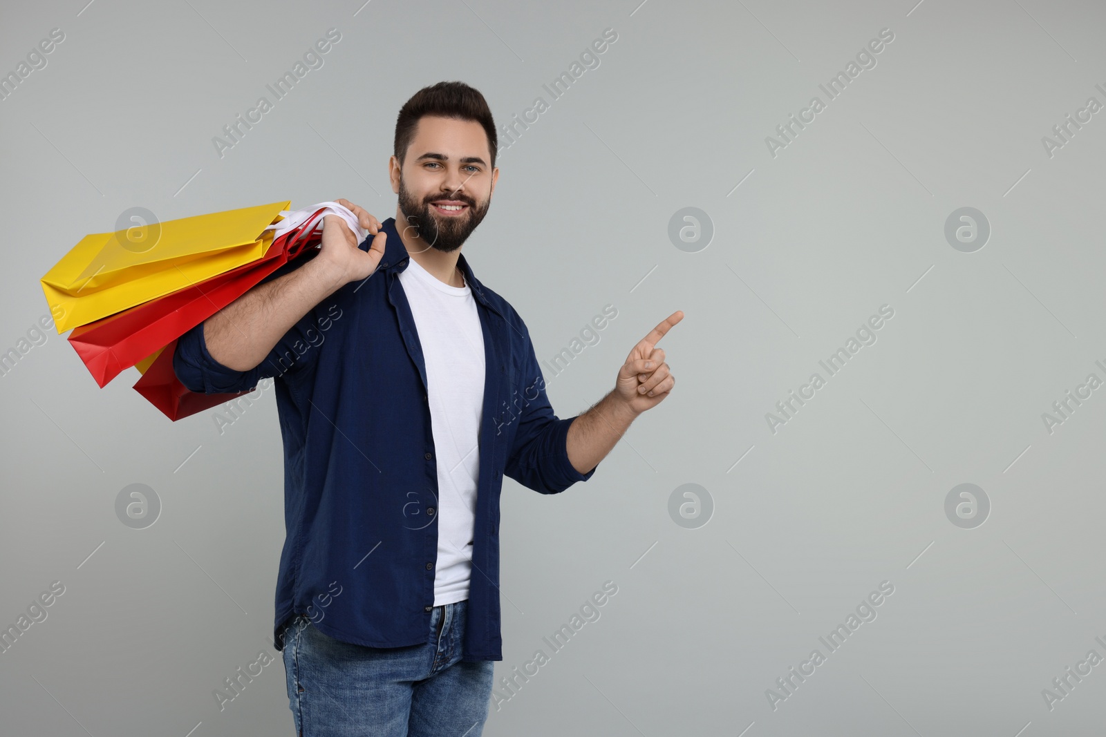 Photo of Happy man with many paper shopping bags pointing at something on grey background. Space for text