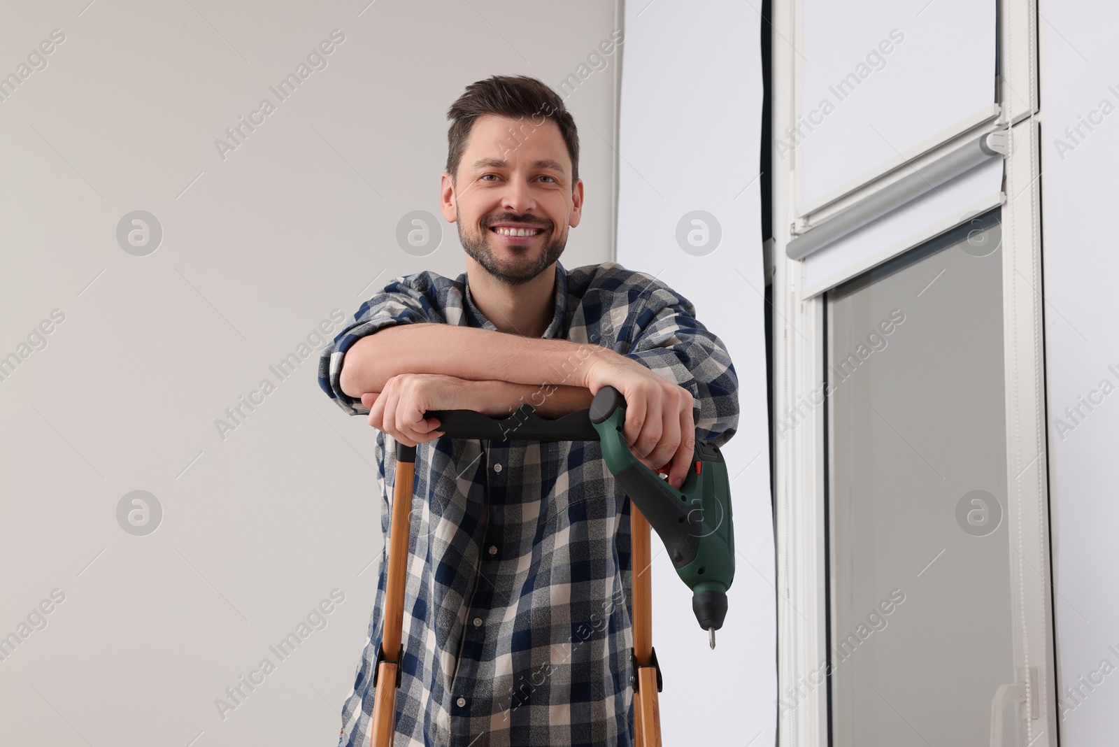 Photo of Man with drill on stepladder near window indoors. Roller blinds installation