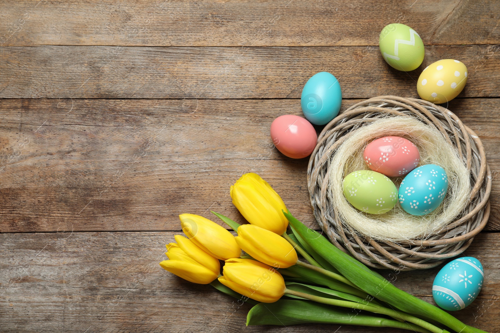 Photo of Flat lay composition of wicker nest with painted Easter eggs and tulips on wooden table, space for text