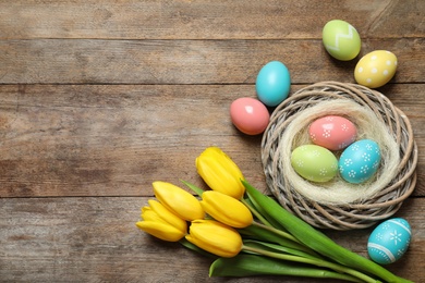 Photo of Flat lay composition of wicker nest with painted Easter eggs and tulips on wooden table, space for text