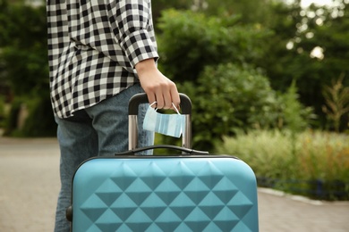 Photo of Woman with suitcase and protective mask outdoors, closeup. Travelling during coronavirus pandemic