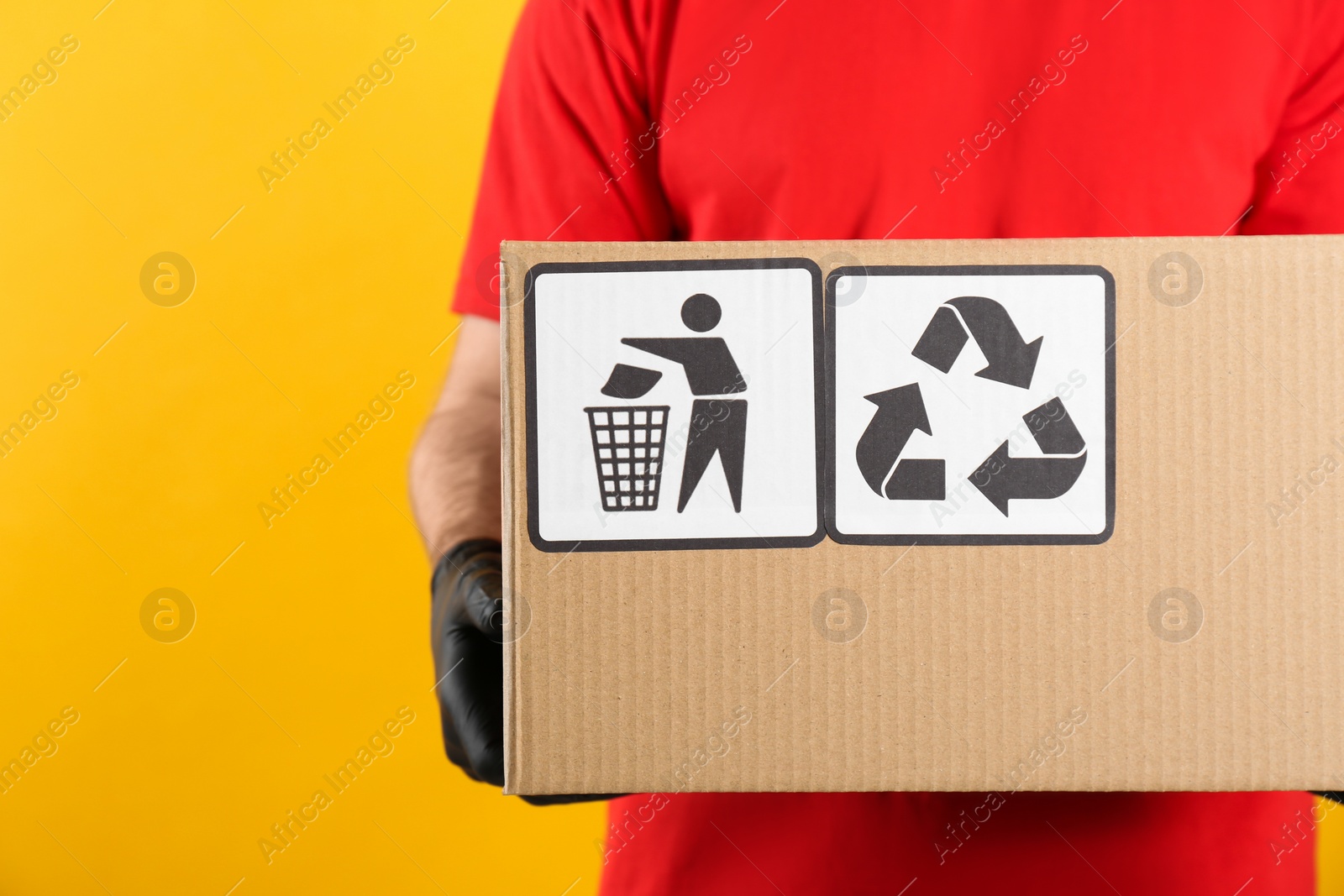 Photo of Courier holding cardboard box with different packaging symbols on yellow background, closeup. Parcel delivery