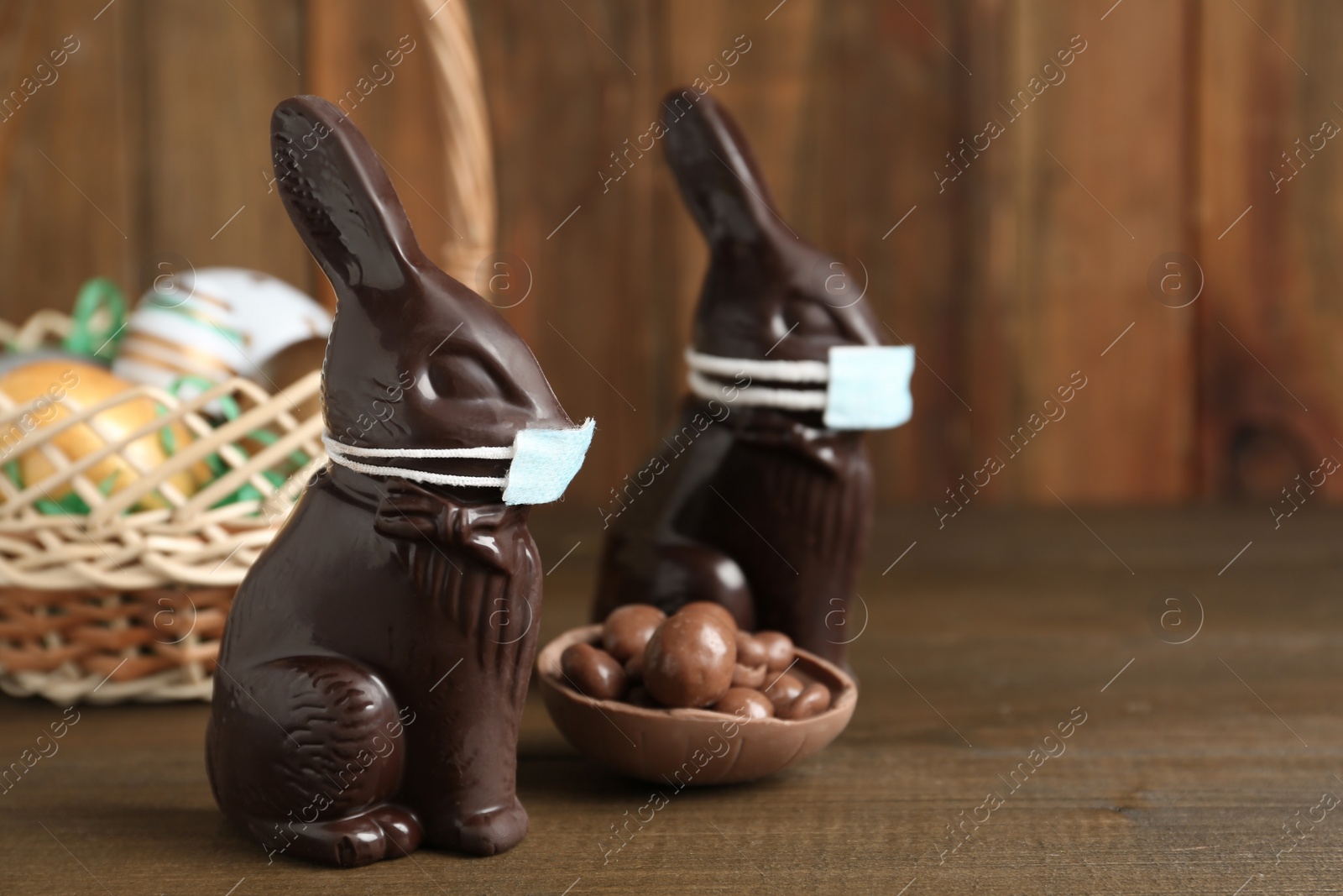 Photo of Chocolate bunnies with protective masks and eggs on wooden table, space for text. Easter holiday during COVID-19 quarantine