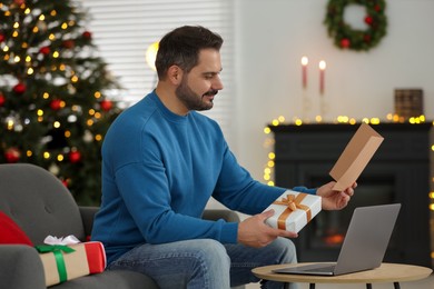 Photo of Celebrating Christmas online with exchanged by mail presents. Man with greeting card and gift box during video call on laptop at home