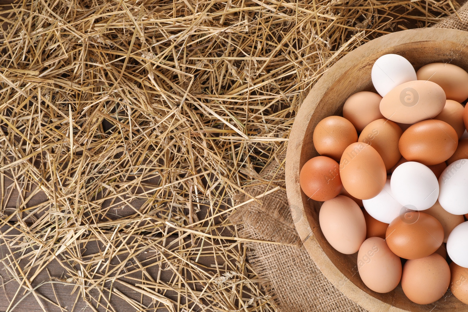 Photo of Fresh chicken eggs in bowl and dried straw on wooden table, top view