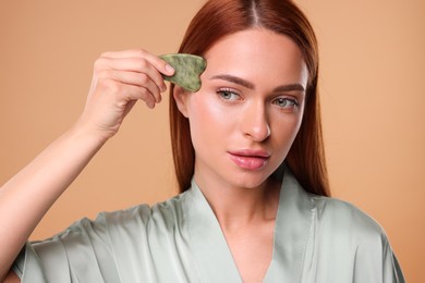 Young woman massaging her face with jade gua sha tool on pale orange background
