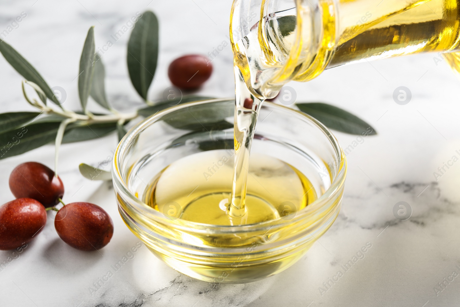 Photo of Pouring with jojoba oil from jug into bowl on white marble table