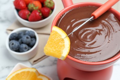 Photo of Fondue pot of melted chocolate and fork with fresh orange on white table, closeup