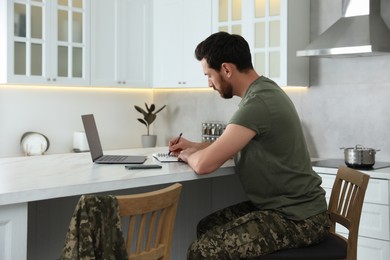 Photo of Soldier taking notes while working with laptop at white marble table in kitchen. Military service