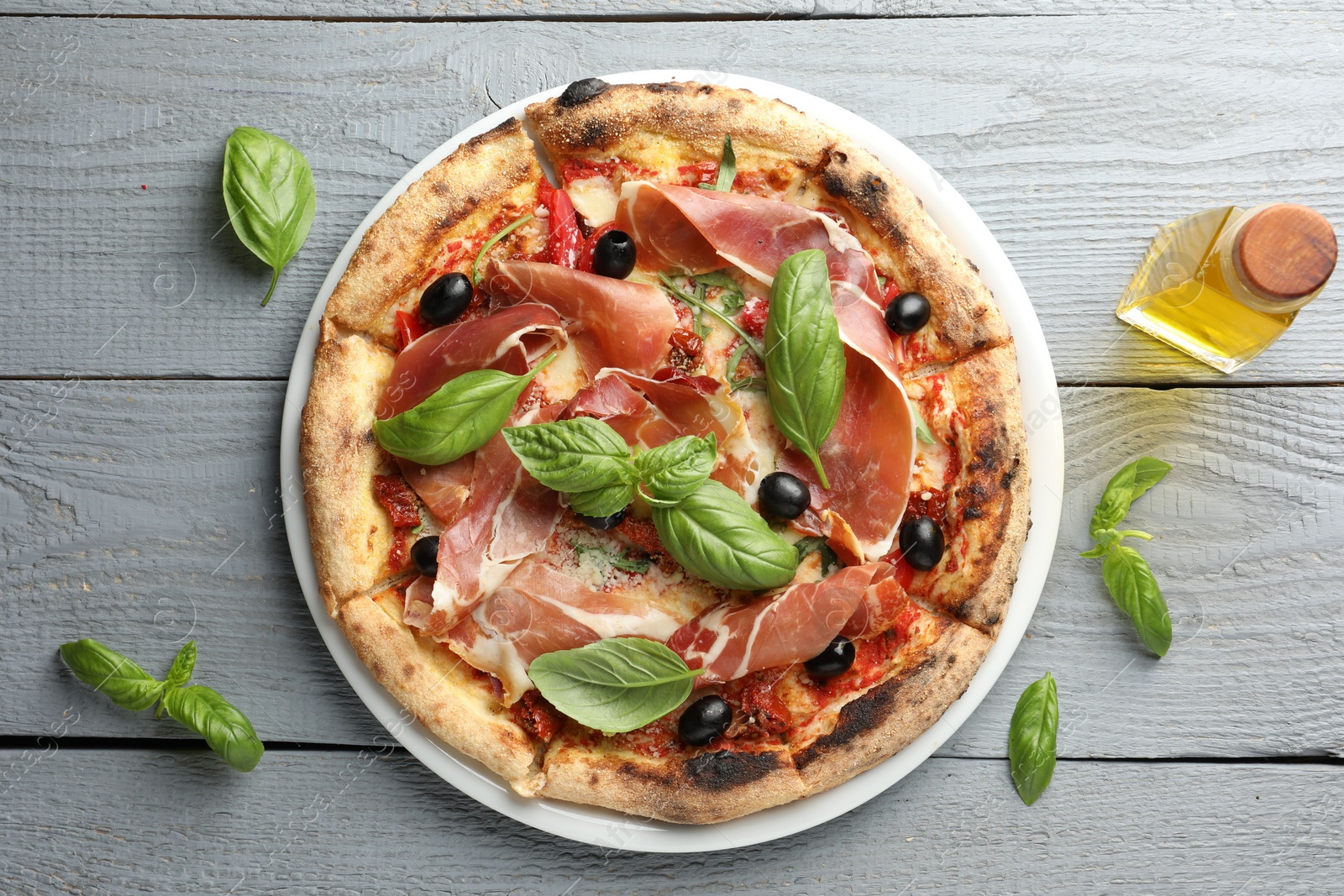 Photo of Tasty pizza with cured ham, olives, sun-dried tomato and basil on gray wooden table, top view