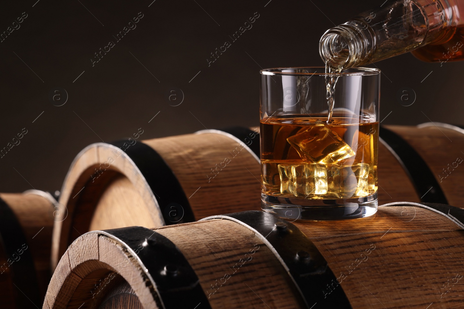 Photo of Pouring whiskey from bottle into glass on wooden barrel against dark background, closeup