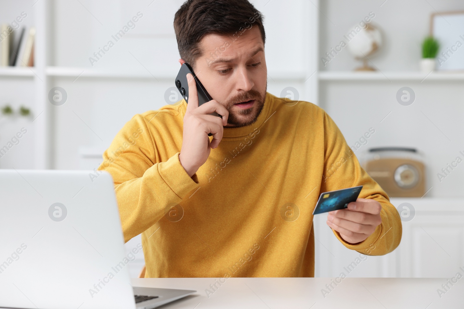 Photo of Upset man with credit card talking on smartphone near laptop at home. Be careful - fraud